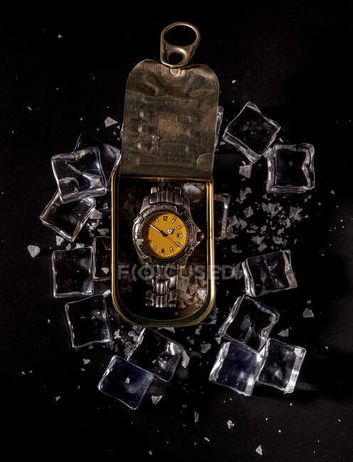 Сверху часы, помещенные в металлическую банку на черной поверхности с разбросанными кубиками льда, демонстрируют концепцию хорошо сохранившегося гаджета — стоковое фото
