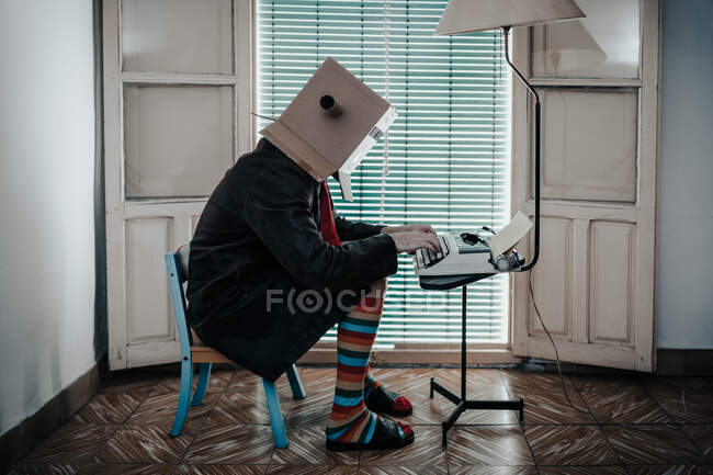 Man wearing cardboard box and striped socks sitting on chair typing on retro typewriter — Stock Photo