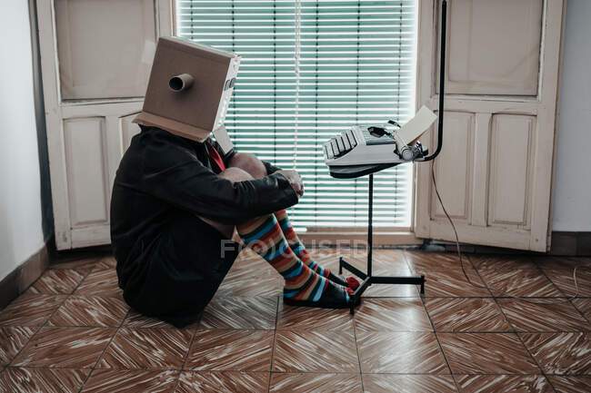 Hombre con caja de cartón y calcetines a rayas sentado en el suelo por silla con máquina de escribir retro - foto de stock