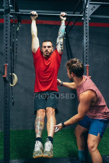 Determined male athlete in sports shorts and t shirt doing exercises on crossbar and pumping muscles under supervision of coach — Stock Photo