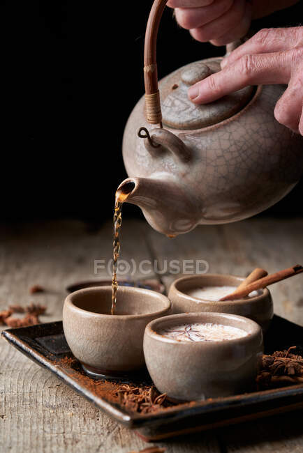 Persona de la cosecha con tetera que vierte Masala chai en cuencos de cerámica colocados en bandeja con anís estrellado y palitos de canela - foto de stock