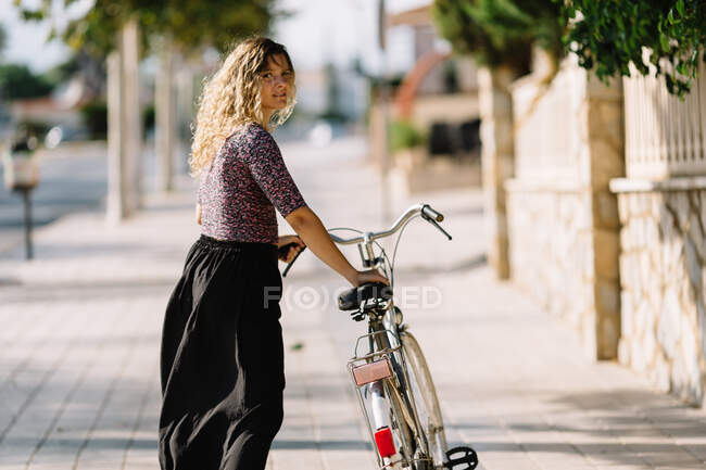 Mujer tranquila en traje de verano caminando con bicicleta en el parque en un día soleado y mirando hacia otro lado - foto de stock