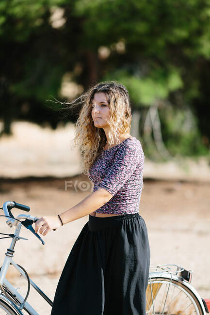 Calma femminile in abito estivo a piedi con bici in parco nella giornata di sole e guardando altrove — Foto stock
