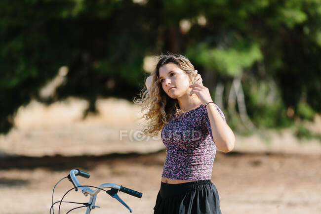 Mujer tranquila en traje de verano caminando con bicicleta en el parque en un día soleado y mirando hacia otro lado - foto de stock