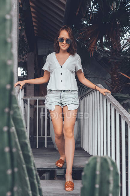Bajo ángulo de mujer delgada feliz en camisa casual gris y pantalones cortos con smartphone tocando gafas de sol y mirando hacia otro lado con interés mientras se apoya en la barandilla en la escalera de madera en el cómodo hotel resort en Bali - foto de stock
