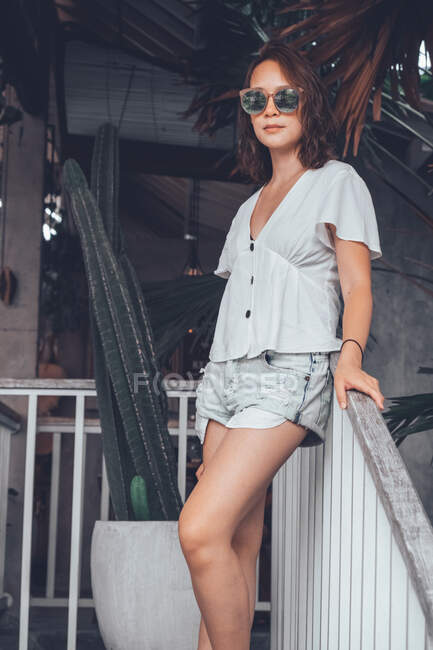 Vista lateral de ángulo bajo de mujer delgada feliz en camisa casual gris y pantalones cortos con smartphone tocando gafas de sol y mirando a la cámara con interés mientras se apoya en la barandilla en la escalera de madera en el cómodo hotel resort en Bali - foto de stock