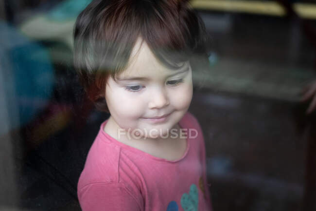 Вид сбоку маленькой девочки, стоящей у окна и выглядывающей, проводя время дома — стоковое фото