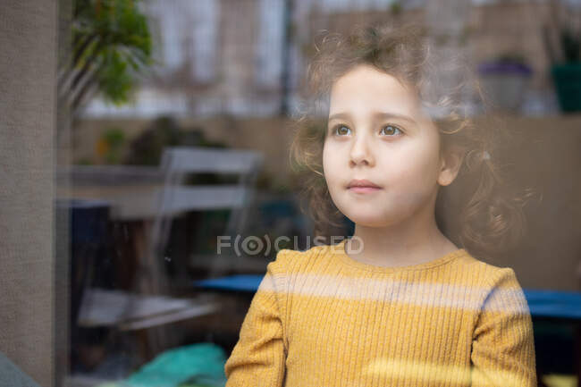 Menina calma com cabelo encaracolado em pé perto da janela e olhando para longe pensativo enquanto passa o tempo em casa e sonhando com aventuras — Fotografia de Stock