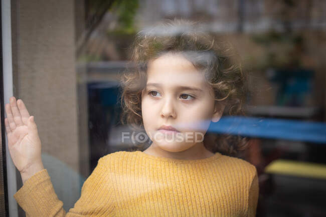 Спокойная маленькая девочка с вьющимися волосами, стоящая у окна и задумчиво глядя в сторону, проводя время дома и мечтая о приключениях — стоковое фото