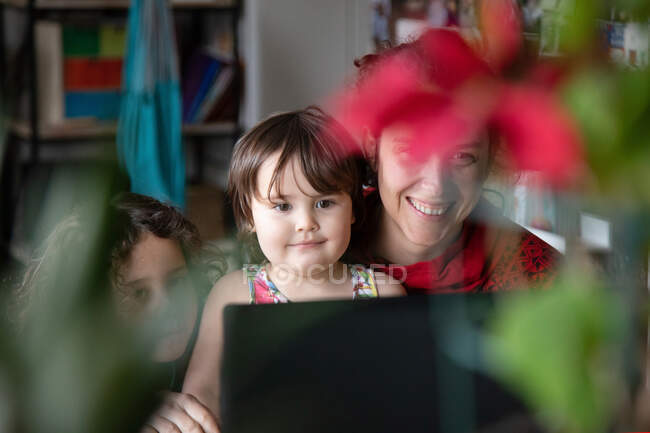 Весела молода жінка з милою маленькою донькою сидить за столом і насолоджується відео розмовою з друзями через ноутбук, проводячи вечір вдома — стокове фото