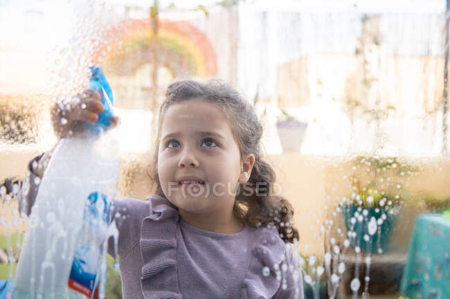 Petite fille positive en robe décontractée avec vaporisateur de verre à laver propre dans la chambre avec arc-en-ciel peint sur la fenêtre pendant la quarantaine de coronavirus — Photo de stock