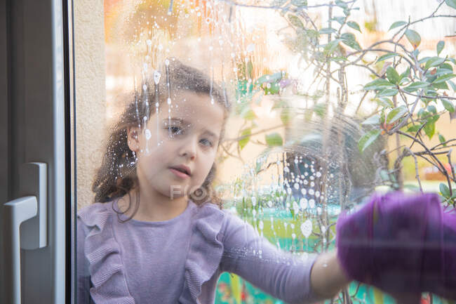 Petite fille positive en robe décontractée avec vaporisateur de verre à laver propre dans la chambre avec arc-en-ciel peint sur la fenêtre pendant la quarantaine de coronavirus — Photo de stock