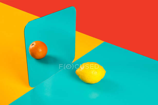 Fondo vibrante con reflejo espejo de naranja fresca como limón sobre superficie azul en composición con áreas rojas y amarillas vacías - foto de stock