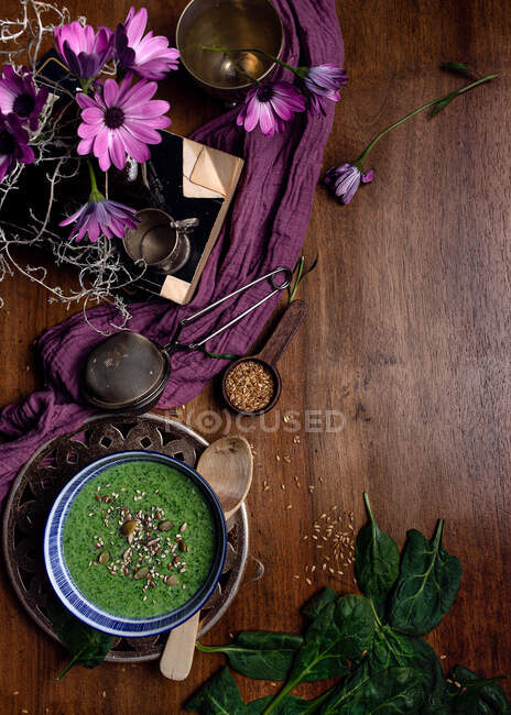 Desde arriba de cuenco de cerámica de batido verde fresco con semillas de calabaza y sésamo en composición con tela delicada púrpura y ramo de flores frescas de margarita violeta en jarrón colocado en mesa de madera marrón - foto de stock