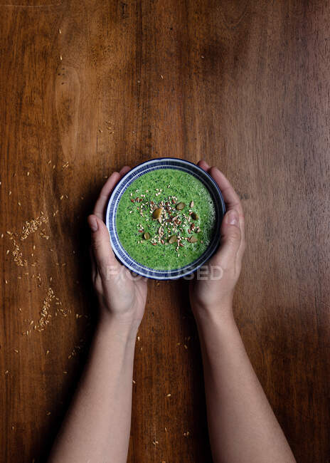 Top view of unrecognizable female holding with both hands ceramic small bowl of fresh green smoothie with adding of pumpkin seeds and sesame against brown wooden table — Stock Photo