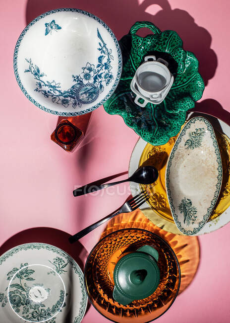 Вид зверху різної форми різнокольоровий керамічний і скляний старовинний посуд в композиції з металевими столовими приборами на яскраво-рожевому фоні в яскравих променях на кухні — стокове фото