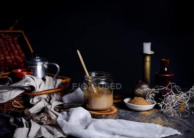 Still life of yummy caramel with cane sugar powder as ingredient placed on table in arrangement with tea set in wicker basket and various decor elements against blurred dark background in studio — Stock Photo