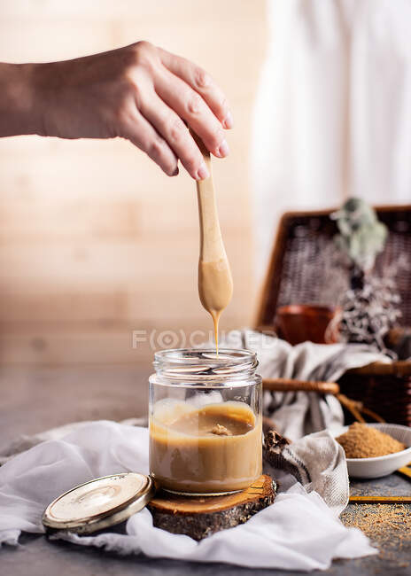 Cocinera femenina irreconocible sosteniendo cucharadita sobre frasco de vidrio transparente de sabroso caramelo colocado en soporte de madera entre tela blanca en la mesa al lado del tazón con azúcar de caña en polvo mientras cocina el postre en casa - foto de stock