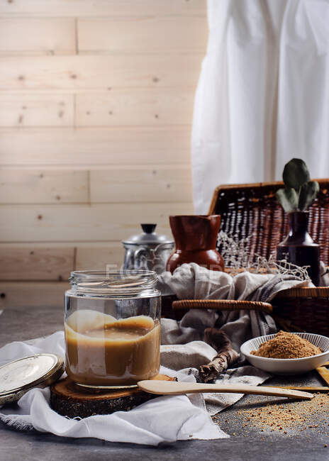 Vaso in vetro trasparente di gustoso caramello posto su basamento in legno tra tessuto bianco sul tavolo accanto alla ciotola con zucchero di canna in polvere a casa — Foto stock