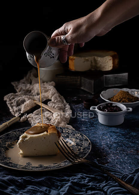 Бездоганна жіноча кухарка наливає карамель на шматок смачного сирного пирога на керамічну пластинку, поміщену на синьому столі на чорному тлі після приготування десерту вдома. — стокове фото