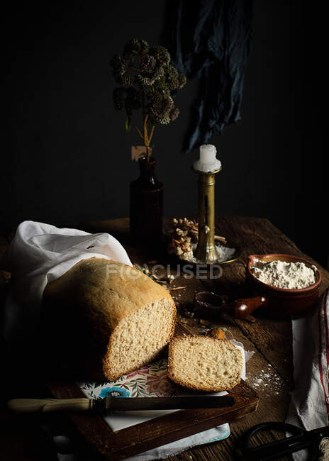 Loaf of homemade bread on cutting board and fresh curd cheese placed on kitchen table with candlestick and vase with plant — Stock Photo