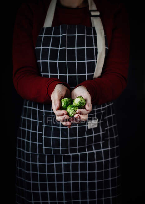 Жіночий кухар у пробному фартусі стоїть з мискою свіжої Брюссельської капусти на чорному тлі на кухні. — стокове фото