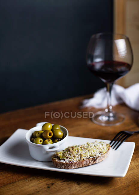 Вид сверху на чашу с маринованными оливками и брускеттой на тарелке, поставленной на стол с бокалом красного вина на кухне — стоковое фото