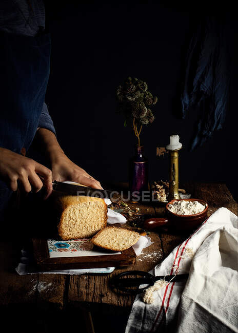 Cropped unrecognizable person wearing apron cutting loaf of homemade bread on wooden board and fresh curd cheese placed on kitchen table with candlestick and vase with plant — Stock Photo