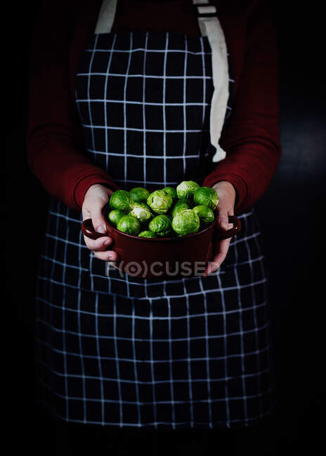 Ernte Köchin in karierter Schürze steht mit Schüssel frischen Rosenkohls auf schwarzem Hintergrund in der Küche — Stockfoto