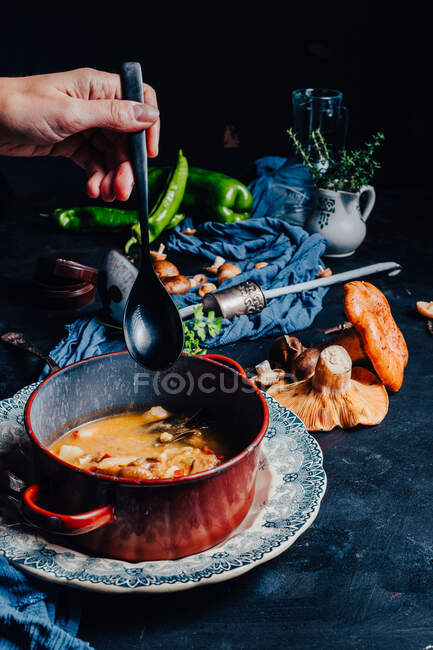 Обрезанная рука неузнаваемого человека, золотая ложка рядом с миской с вкусным супом, поставленным на стол с красными сосновыми грибами и острым зеленым перцем чили, подаваемым на ужин — стоковое фото