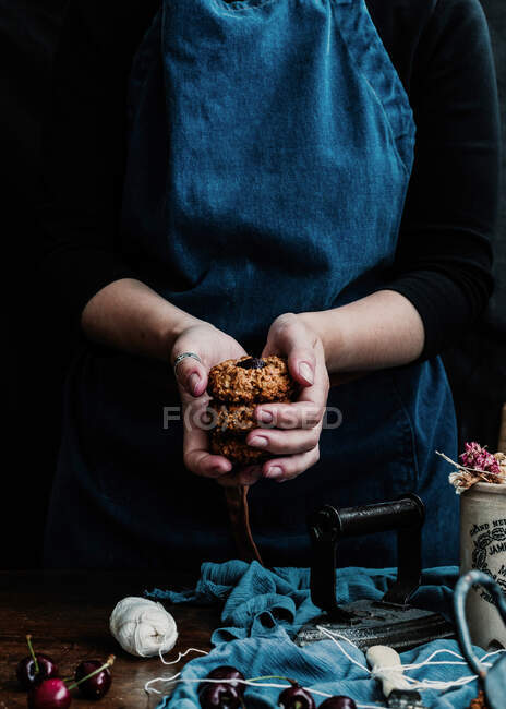 Anonymous woman wearing apron standing with stack of cookies at table with fresh cherry and retro iron on cloth — Stock Photo