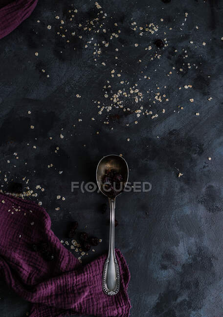 Вид сверху серебряной ложки с сухой черной смородиной, выложенной на стол кухонным инструментом и посыпанной овсянкой — стоковое фото