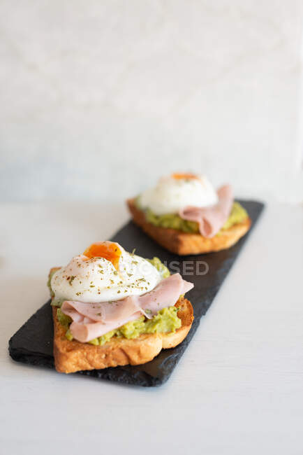Pequeno-almoço saboroso de torradas crocantes decoradas com guacamole e ovos fritos com presunto na cozinha brilhante — Fotografia de Stock