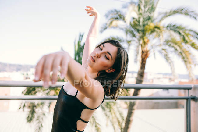 Bailarina femenina elegante en el momento de realizar el elemento con los brazos extendidos mirando hacia fuera en la terraza de verano en el fondo de las palmeras - foto de stock