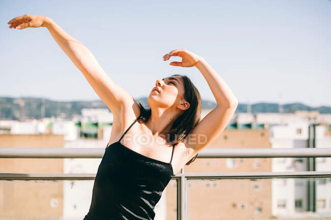Bailarina femenina elegante en el momento de realizar elemento con brazos extendidos mirando hacia arriba en la terraza de verano sobre fondo de palmeras - foto de stock