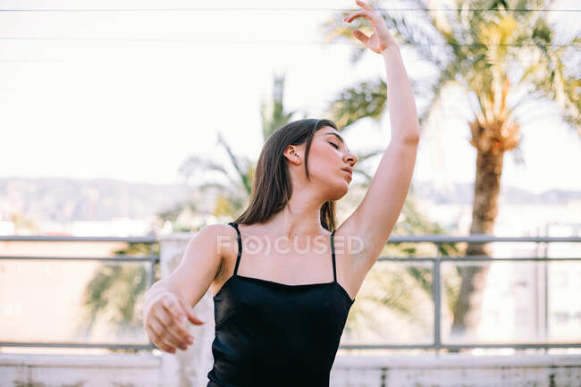 Витончена жіноча танцівниця в момент виконання елемента з витягнутими руками і закритими очима на літній терасі на фоні пальмових дерев — стокове фото