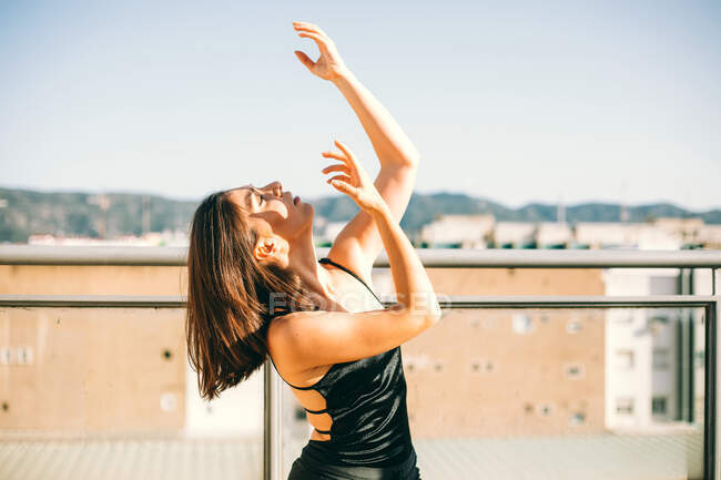 Bailarina femenina elegante en el momento de realizar elemento con brazos extendidos mirando hacia arriba en la terraza de verano sobre fondo de palmeras - foto de stock