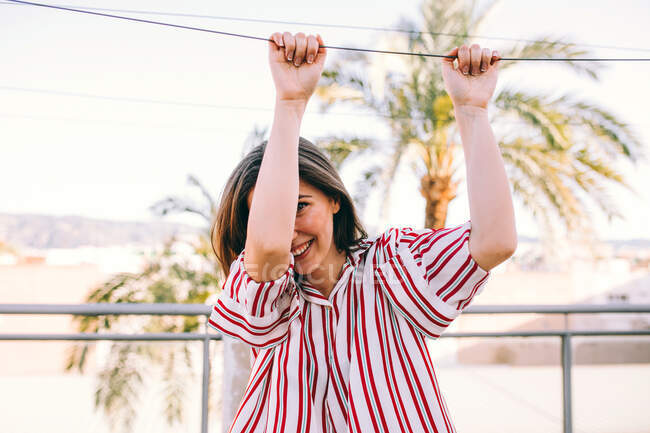Вид сбоку играющей женщины в стильной одежде, держащейся на летней террасе на фоне размытых пальм и смотрящей в камеру — стоковое фото
