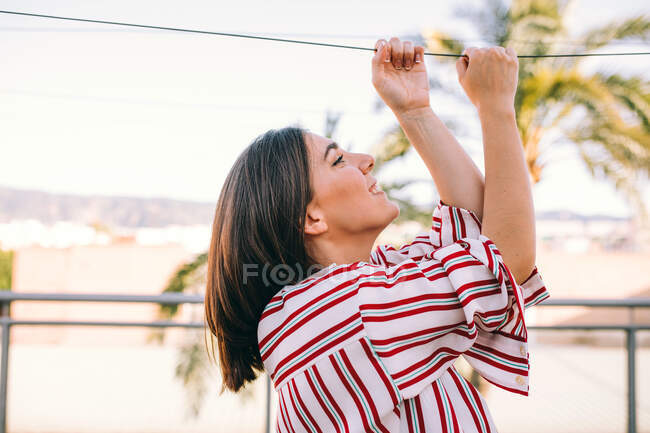 Vista lateral da fêmea brincalhão vestindo roupas elegantes segurando corda no terraço de verão no fundo de palmeiras borradas e olhando para cima — Fotografia de Stock