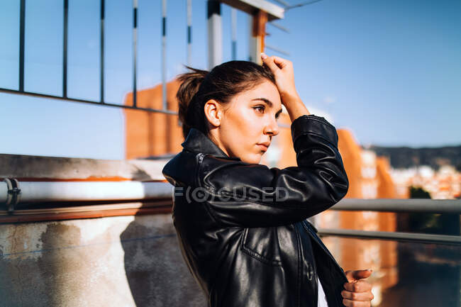 Vista lateral da mulher serena em jaqueta de couro na moda em pé no terraço de verão olhando para longe e desfrutando de tempo ensolarado — Fotografia de Stock