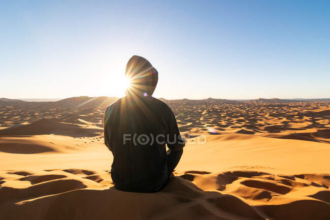 Обратный вид на неузнаваемого туриста, сидящего на песчаной дюне и любующегося величественными пейзажами заката в пустыне в Марокко — стоковое фото