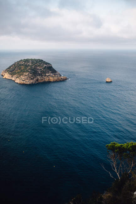 Aerial view of rocky coastline with islet in the middle of the sea close to sea bay with calm blue water against cloudy sky and horizon in light haze at daytime — Stock Photo