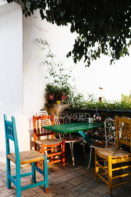 Ensemble coloré de table en bois avec différentes chaises placées près d'une clôture métallique avec des plantes en pot couvertes de lierre sur la véranda de la maison de campagne pendant la journée ensoleillée d'été — Photo de stock