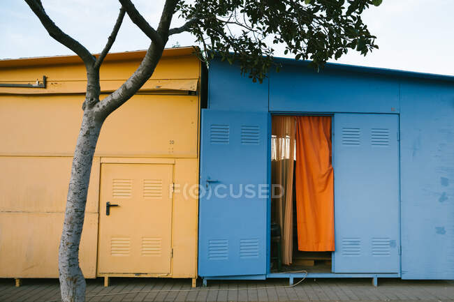Maisons métalliques jaunes et bleues abîmées situées sur le trottoir derrière un arbre vert en zone urbaine pendant la journée ensoleillée d'été avec un ciel bleu clair — Photo de stock