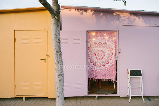 Shabby resistió casas metálicas amarillas y rosadas ubicadas en el pavimento detrás de un árbol en el área urbana durante el soleado día de verano con cielo azul claro - foto de stock
