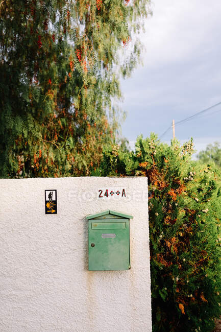 Ржавая стена со старым металлическим почтовым ящиком и номером дома в окружении ветвей деревьев с цветущими красными цветами под облачным небом днем — стоковое фото