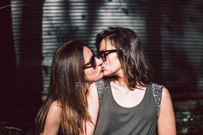 Teneri amanti delle donne che indossano abiti casual e occhiali da sole baciarsi mentre camminano fuori sullo sfondo della strada offuscata nella soleggiata giornata estiva — Foto stock