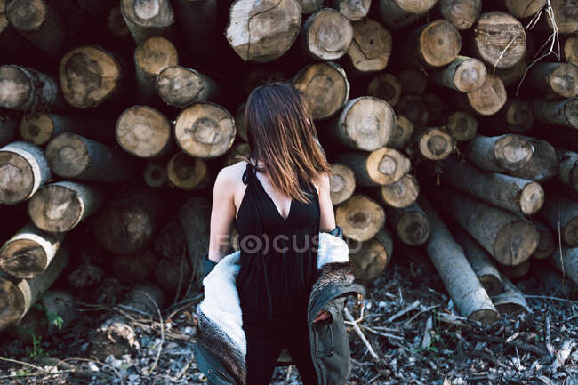 Enigma-Weibchen in schwarzem Freizeitoutfit und heruntergezogenem Pelzparka, die allein an der Wand aus Baumstämmen stehen, mit verstecktem Gesicht — Stockfoto