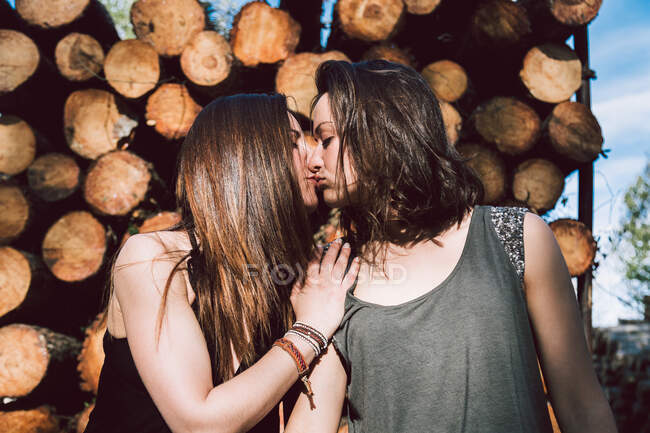Teneri amanti delle donne che indossano abiti casual baciarsi con gli occhi chiusi mentre in piedi contro il muro con i tronchi nella soleggiata giornata estiva — Foto stock