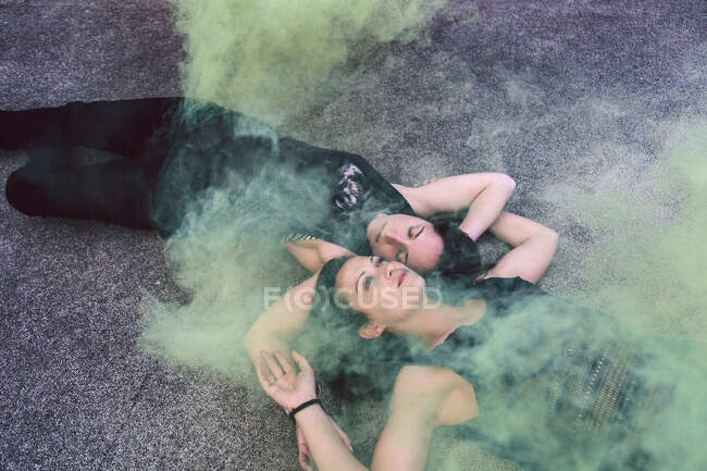 From above of dreamy female lovers in casual clothes lying on asphalt ground head to head and crossing arms under yellow smoke — Stock Photo